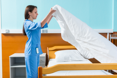 Young nurse in blue unifrom changing bedsheets of hospital bed in the hospital ward